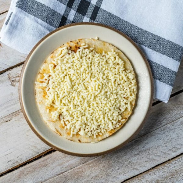 A round, flatbread-like dish covered with shredded white cheese on a light plate, with a checkered napkin.