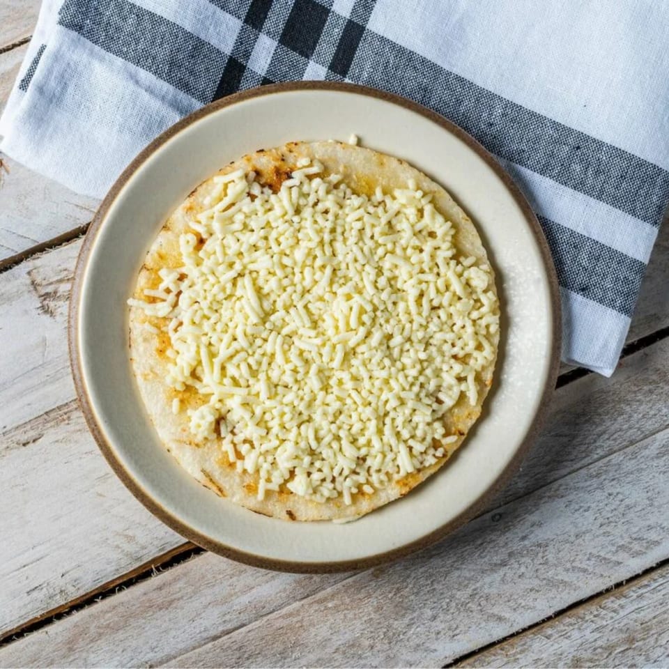A round, flatbread-like dish covered with shredded white cheese on a light plate, with a checkered napkin.