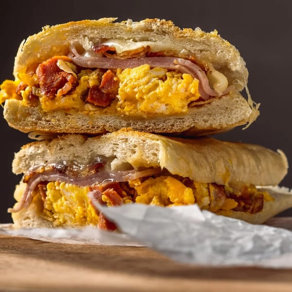 Two stacked breakfast sandwiches with scrambled eggs, ham, bacon, and melted cheese on ciabatta bread.