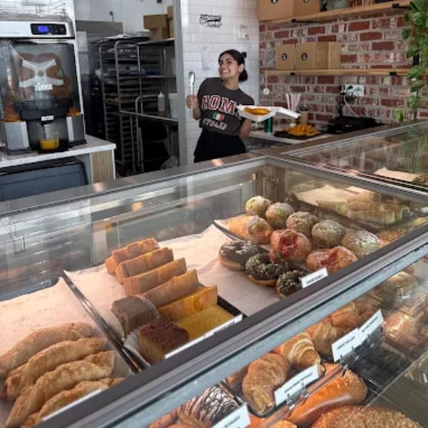 A smiling woman behind a bakery counter filled with pastries, cakes, and donuts.