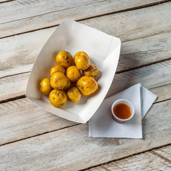 Golden-brown roasted potatoes in a white paper tray, with a side of dipping sauce on a wooden table.