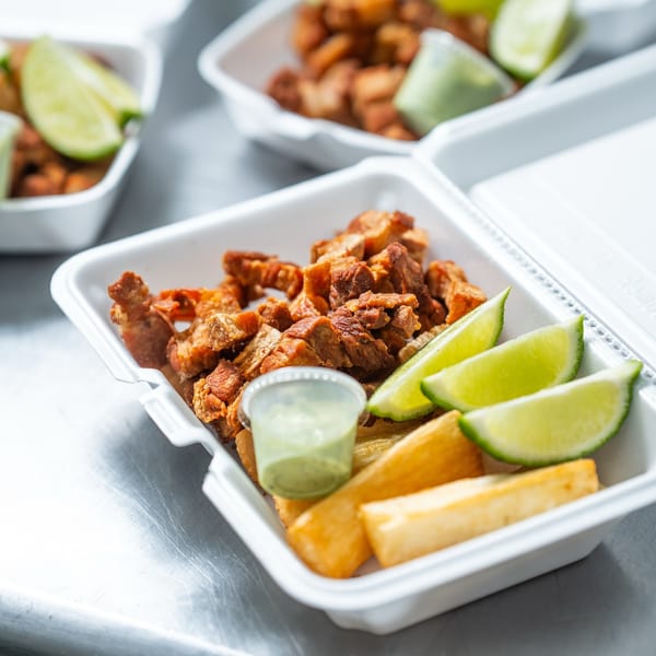 Crispy fried pork chunks, yuca fries, lime wedges, and green sauce in a white takeout container.