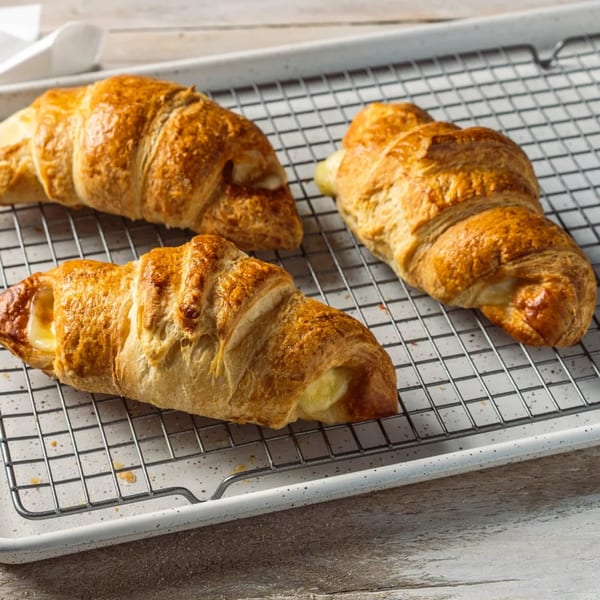 Three golden-brown croissants with creamy filling rest on a cooling rack over a white speckled tray.