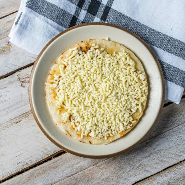 A round, flat arepa covered with shredded white cheese on a plate, next to a checkered napkin.