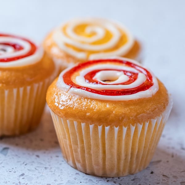 Three glazed cupcakes with swirled red and yellow icing on a white speckled surface.