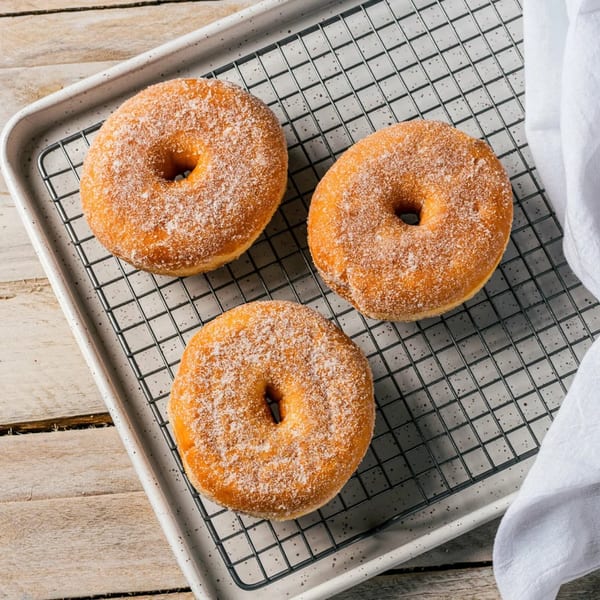 Three sugar-dusted donuts rest on a cooling rack set inside a white tray on a wooden surface.