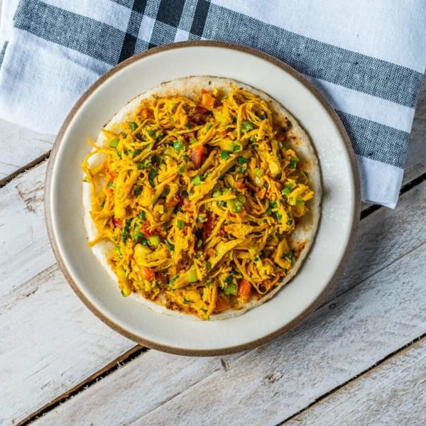 A white plate holds a round flatbread topped with shredded yellow chicken, green onions, and red peppers.