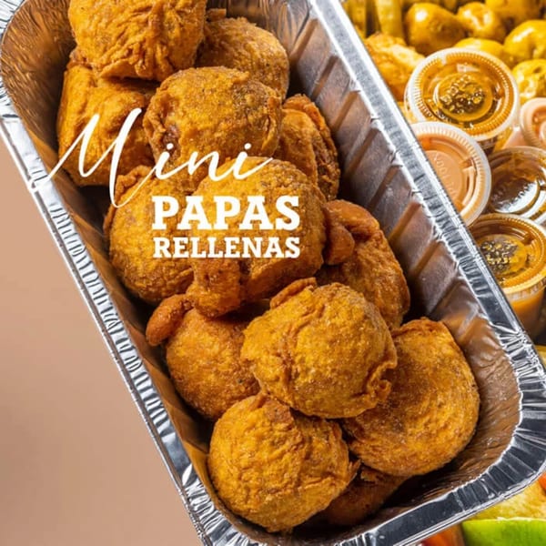 Close-up of golden-brown mini papas rellenas in a foil tray, with sauce containers in the background.